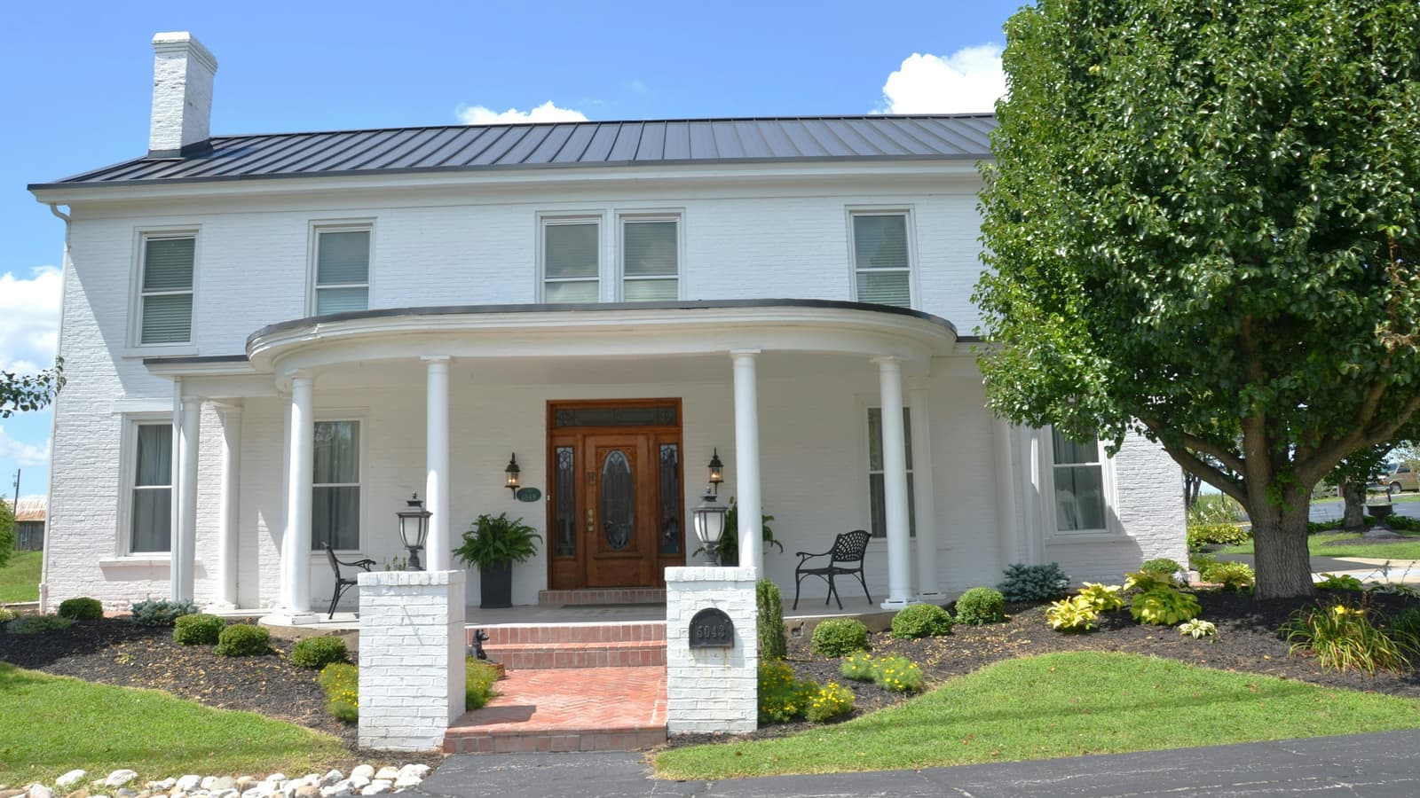 White colonial-style house with a curved porch, wood front door and tidy landscaping under a blue sky